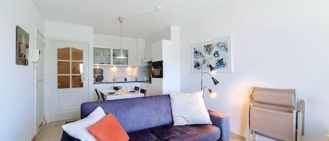Una sala de estar moderna con un sofá azul oscuro, una pequeña mesa con una vela y una cocina al fondo.