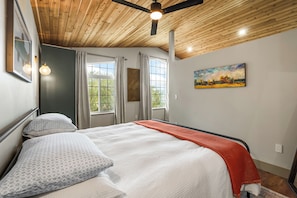 A bedroom with a bed, a ceiling fan, a painting, and windows.