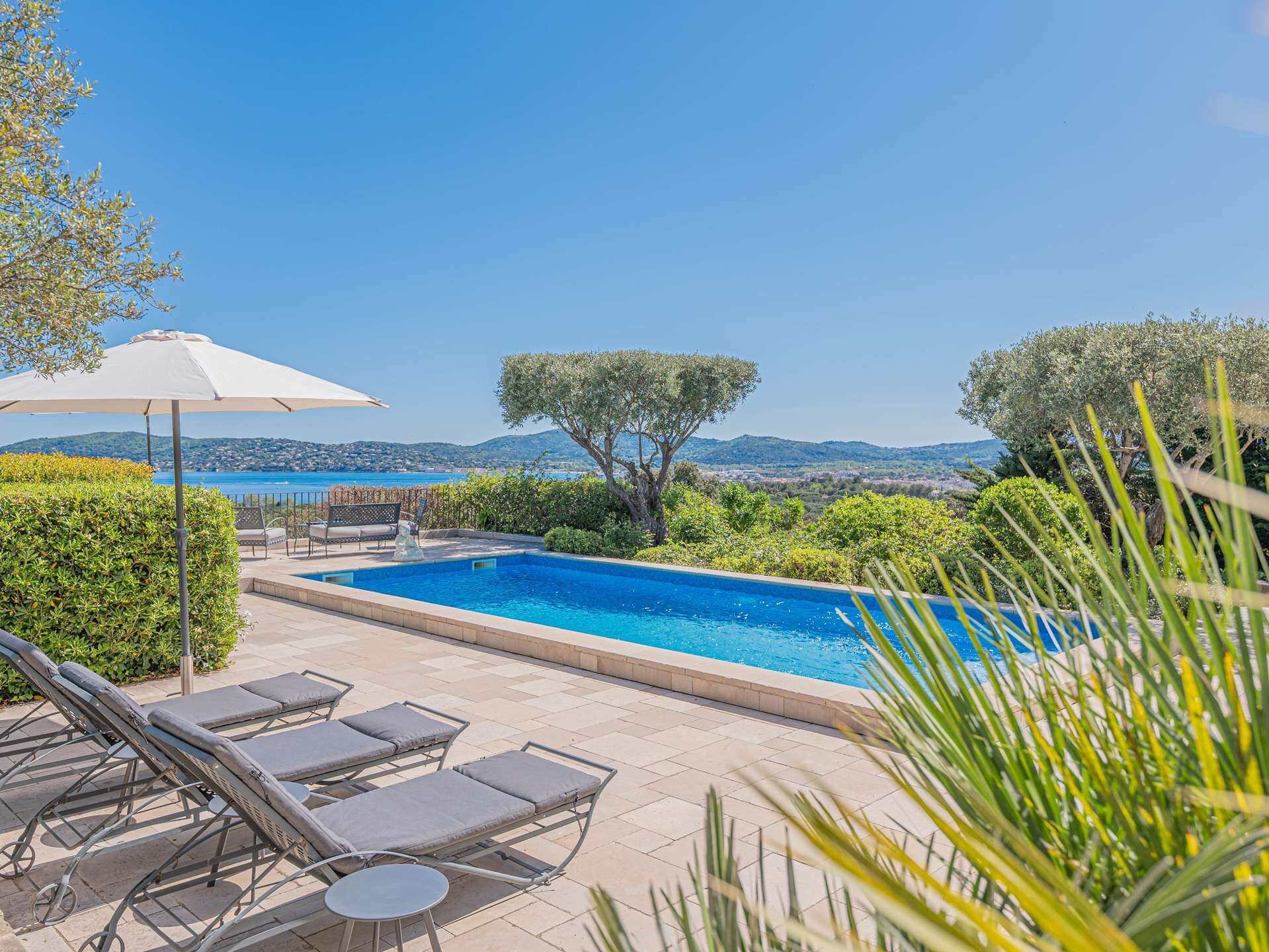 A swimming pool with lounge chairs and a table, surrounded by greenery and a view of the sea.