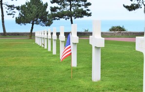 A row of white crosses on a grassy field with a flag in front. - Authentique proche plage D-Day (COURSEULLES-SUR-MER)