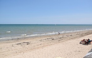 Una playa con arena, un velero a lo lejos y una persona caminando por la orilla.