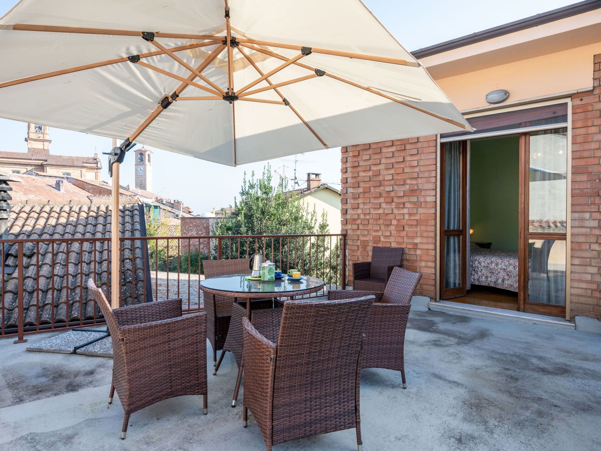 Een terras met een ronde tafel, vier rieten stoelen en een grote parasol.