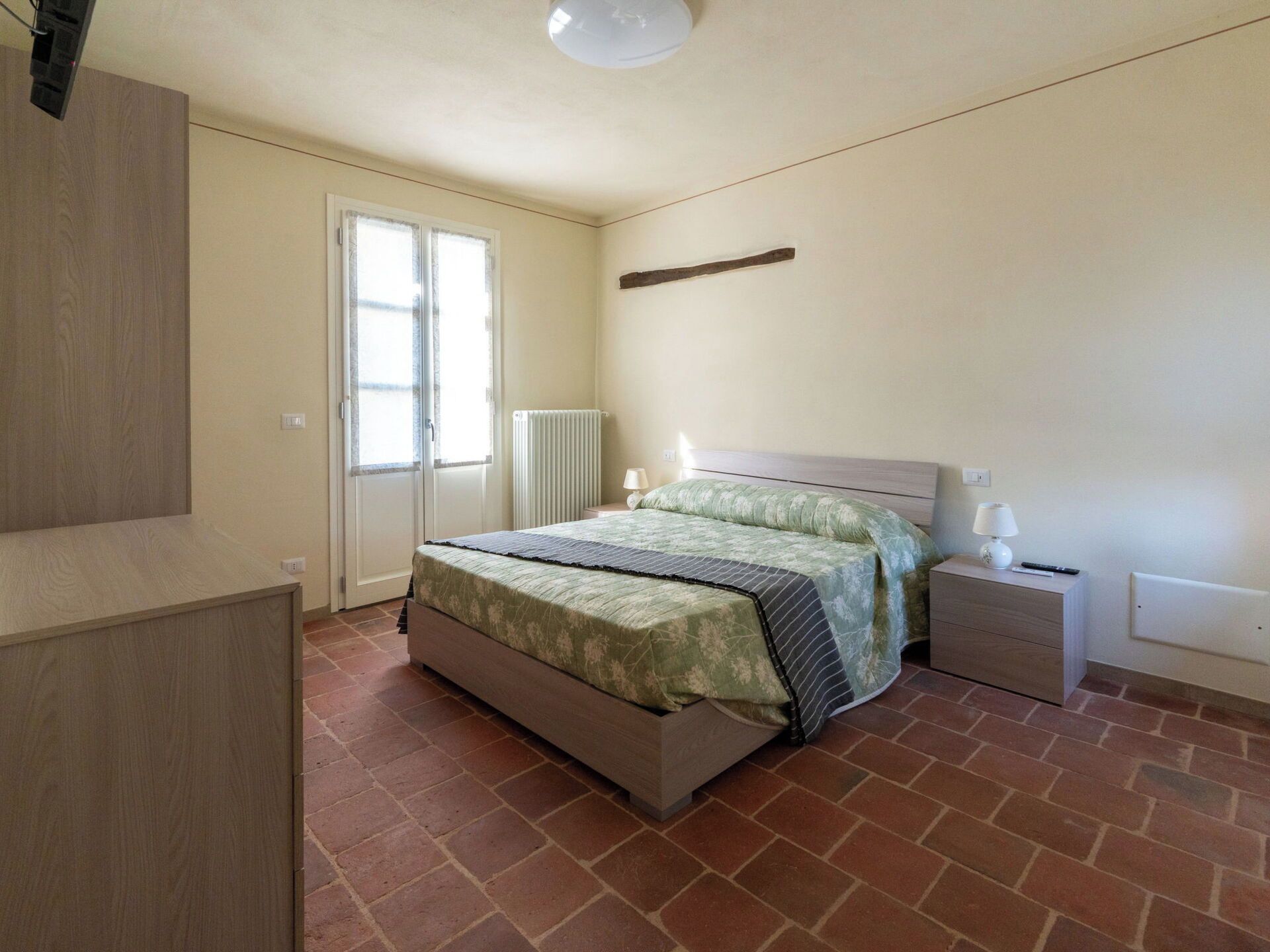Quarto com cama, mesa de cabeceira, radiador e porta com janela.