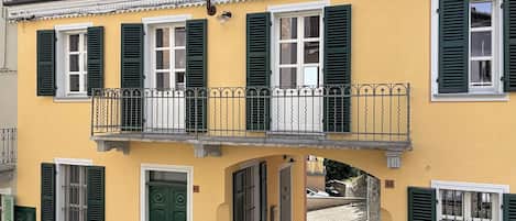 A yellow building with green shutters, a balcony with a metal railing, and a cobblestone street.