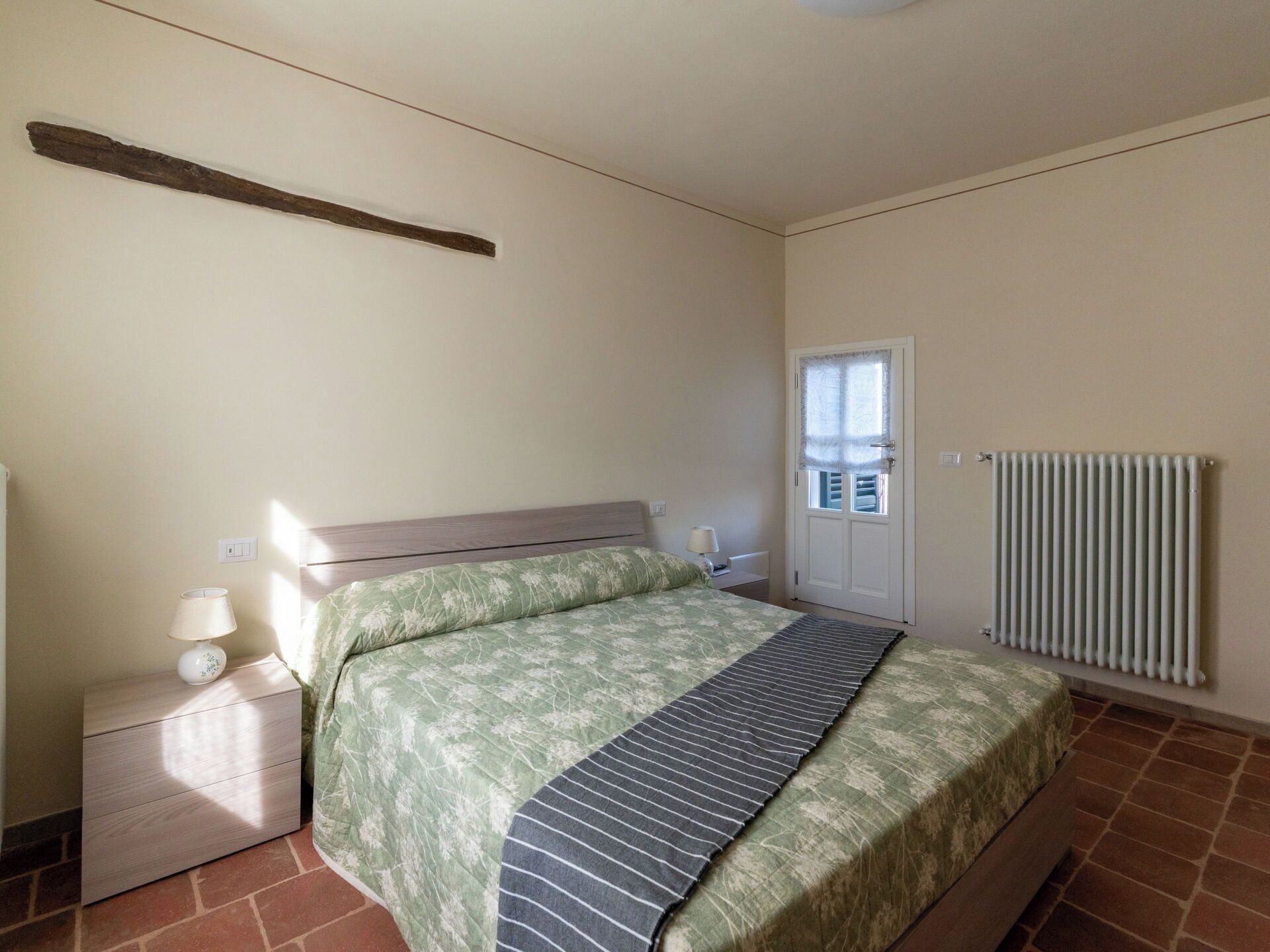 Una camera da letto con un letto, un comodino, una lampada, una finestra e un termosifone.