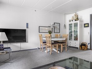 A modern living room with a flat-screen TV, a dining table with chairs, a glass-top coffee table, a cabinet with glass doors, and a wicker basket.