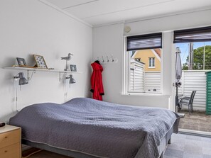 A bedroom with a bed, a nightstand, a wall-mounted shelf with decorative items, a window with blinds, and a view of the outside.