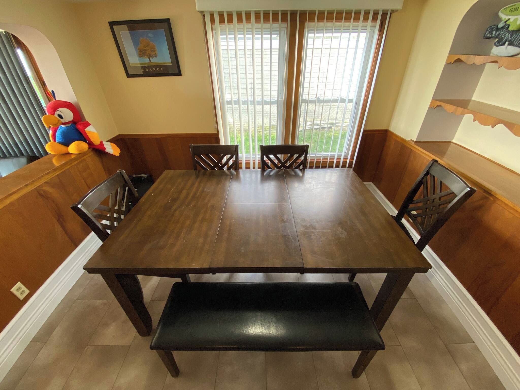 Comedor con mesa de madera, cuatro sillas, un banco y un amplio ventanal con persianas.
