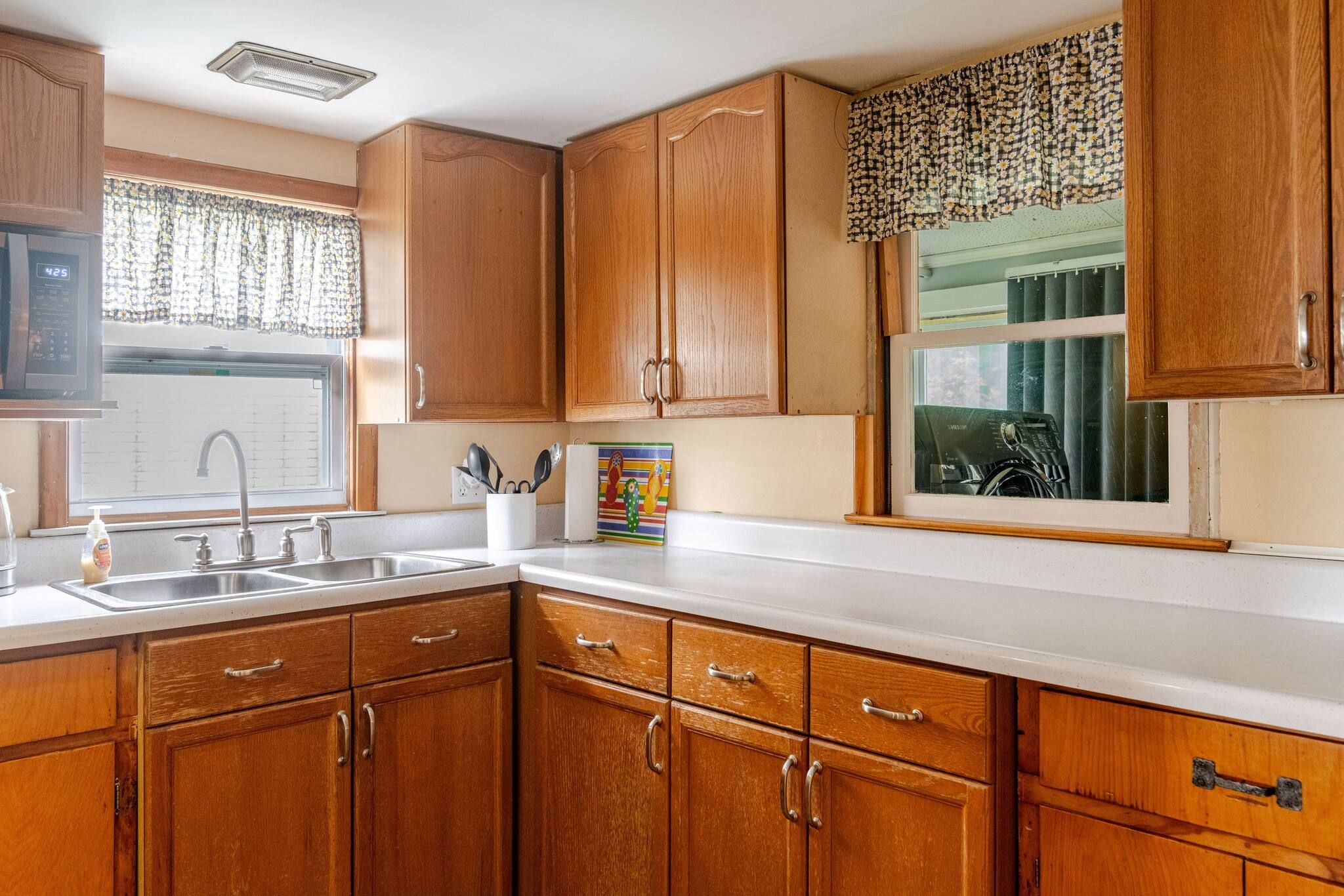 A kitchen with wooden cabinets, a white countertop, a sink, and a window with a patterned curtain.