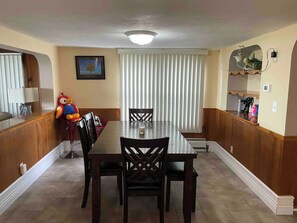 Un coin repas avec une table et des chaises, une fenêtre avec des stores, une étagère fixée au mur et un tableau encadré.