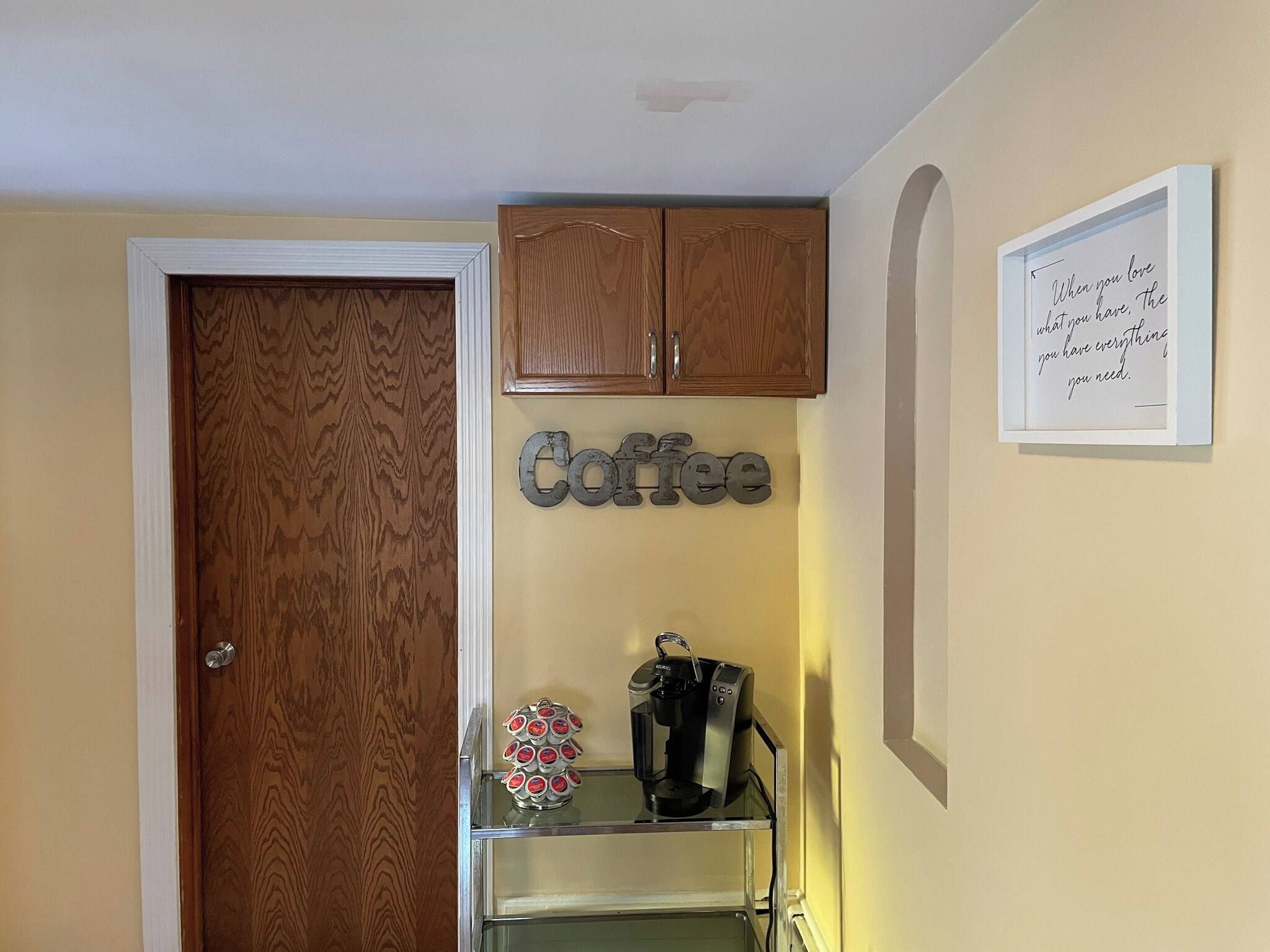 Una habitación con puerta de madera, decoración de temática de café en la pared, una cafetera sobre una estantería y una cita motivacional en la pared.