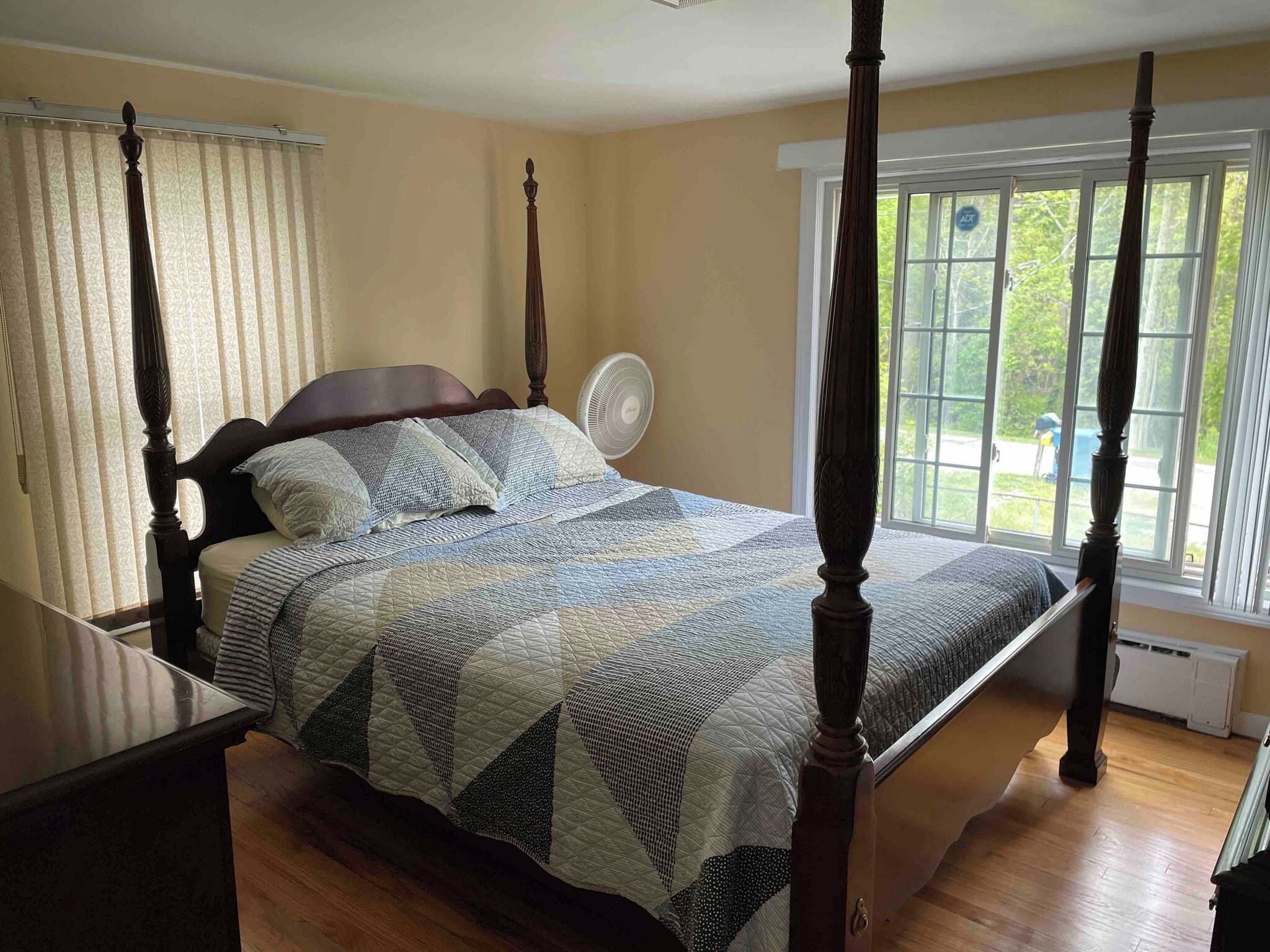 A four-poster bed with a quilt, a standing fan, and a window with blinds.