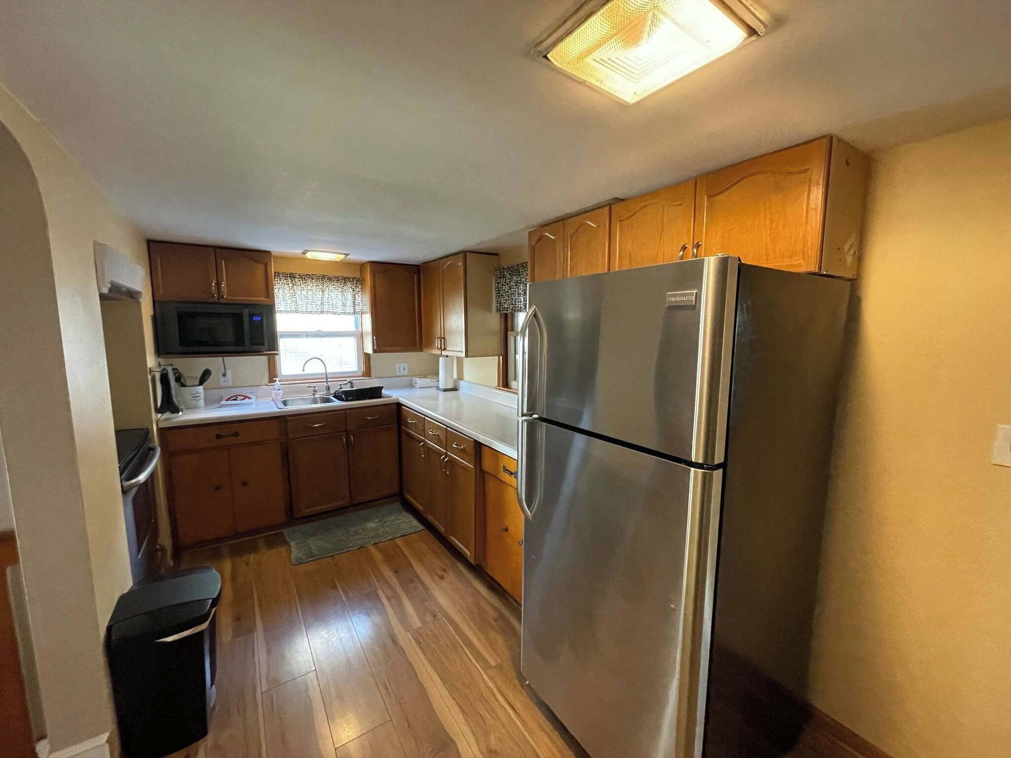 A compact kitchen with wooden cabinets, a stainless steel refrigerator, a microwave, and a window with patterned curtains.