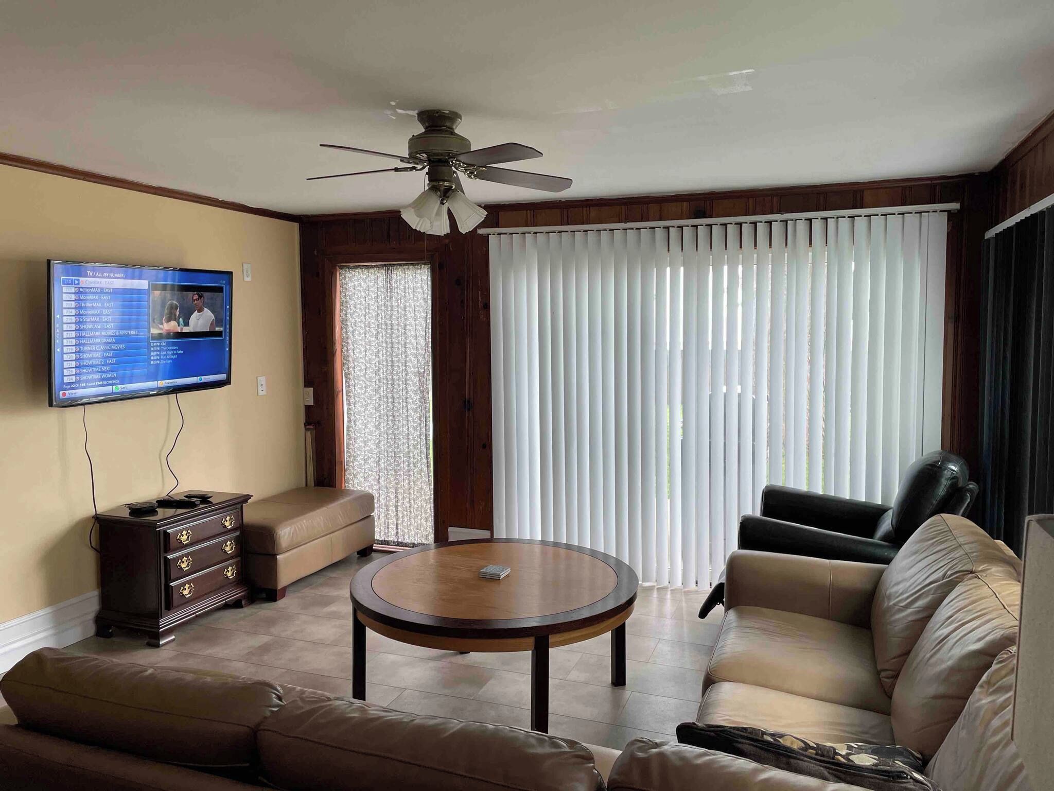 A living room with a TV mounted on the wall, a ceiling fan, a sofa, a coffee table, and a window with blinds.