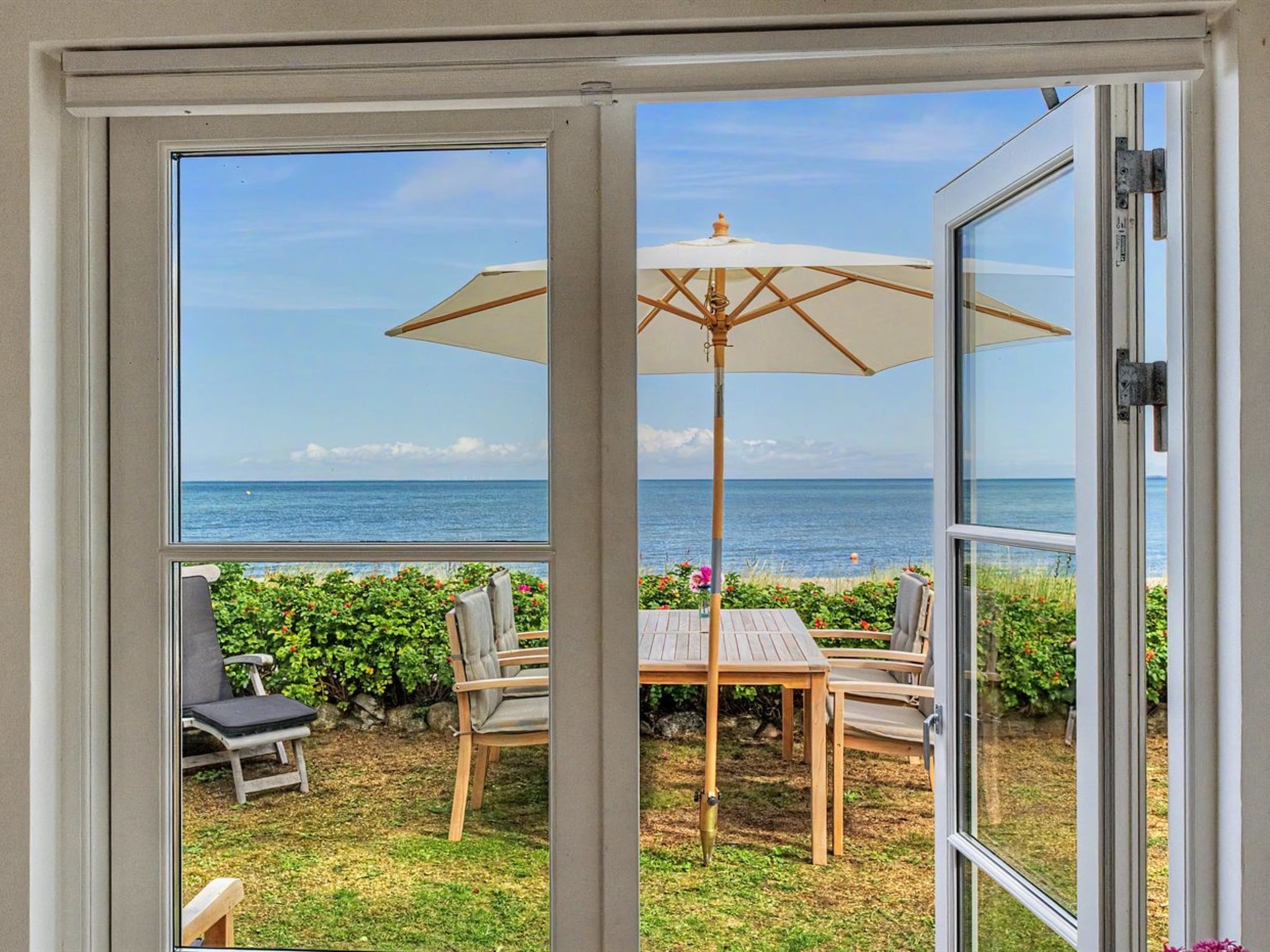 A glass door leading to an outdoor patio with a table, chairs, and an umbrella, overlooking a beach.