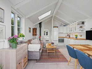 Eine moderne Küche mit weißen Schränken, ein Essbereich mit einem Holztisch und blauen Stühlen sowie ein Wohnzimmer mit einer Couch und einem Couchtisch.