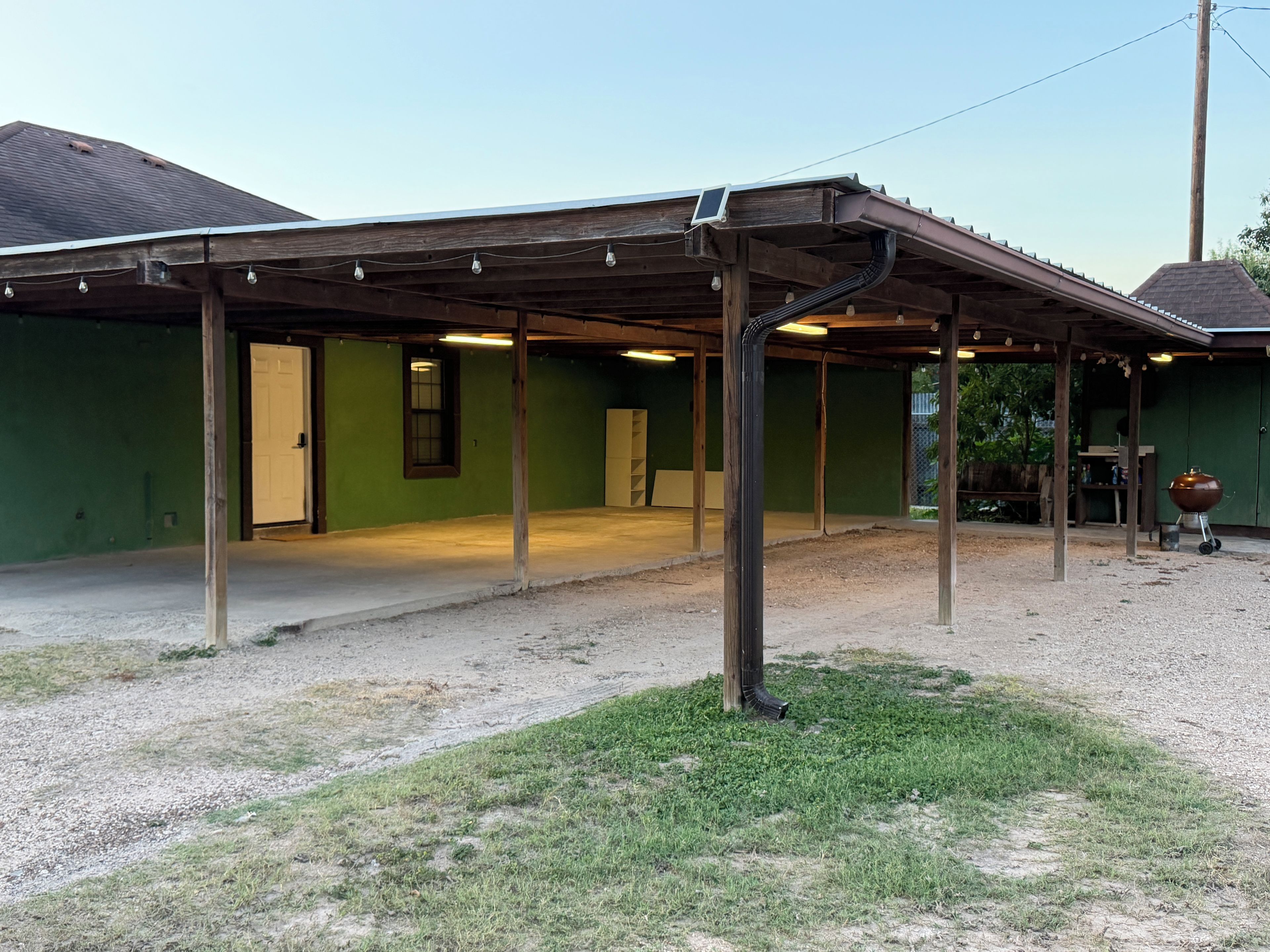 Un espacio al aire libre techado con suelo de grava, una estructura de madera y un edificio verde al fondo.