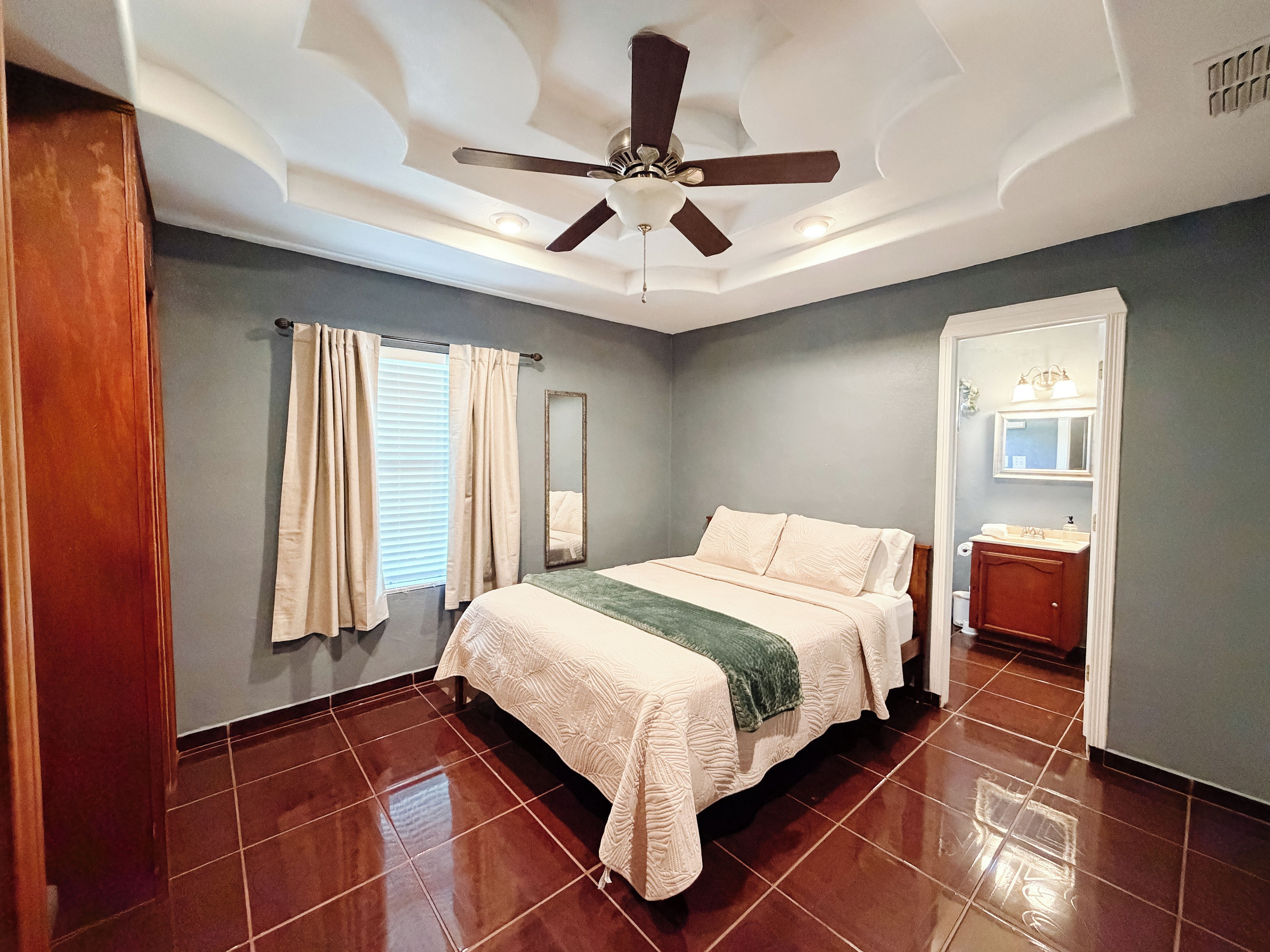 A bedroom with a bed, a ceiling fan, a window with curtains, a mirror, and a bathroom with a sink and toilet.