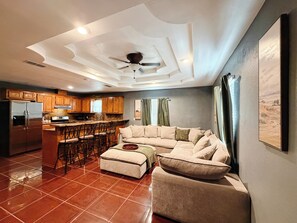 Een moderne keuken met een centraal eiland, een eethoek met barstoelen en een woonkamer met een grote bank en voetenbank.