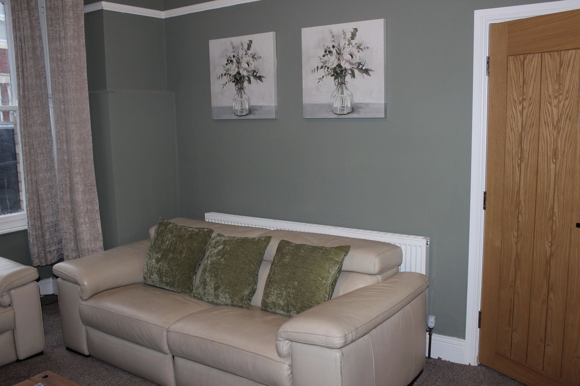 A living room with a beige sofa, two cushions, a radiator, and two wall-mounted paintings of flowers.
