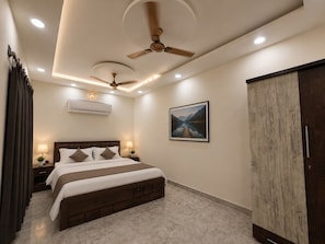 Quarto de hotel com cama, ventiladores de teto, iluminação embutida, ar condicionado, pintura emoldurada de paisagem e guarda-roupa.