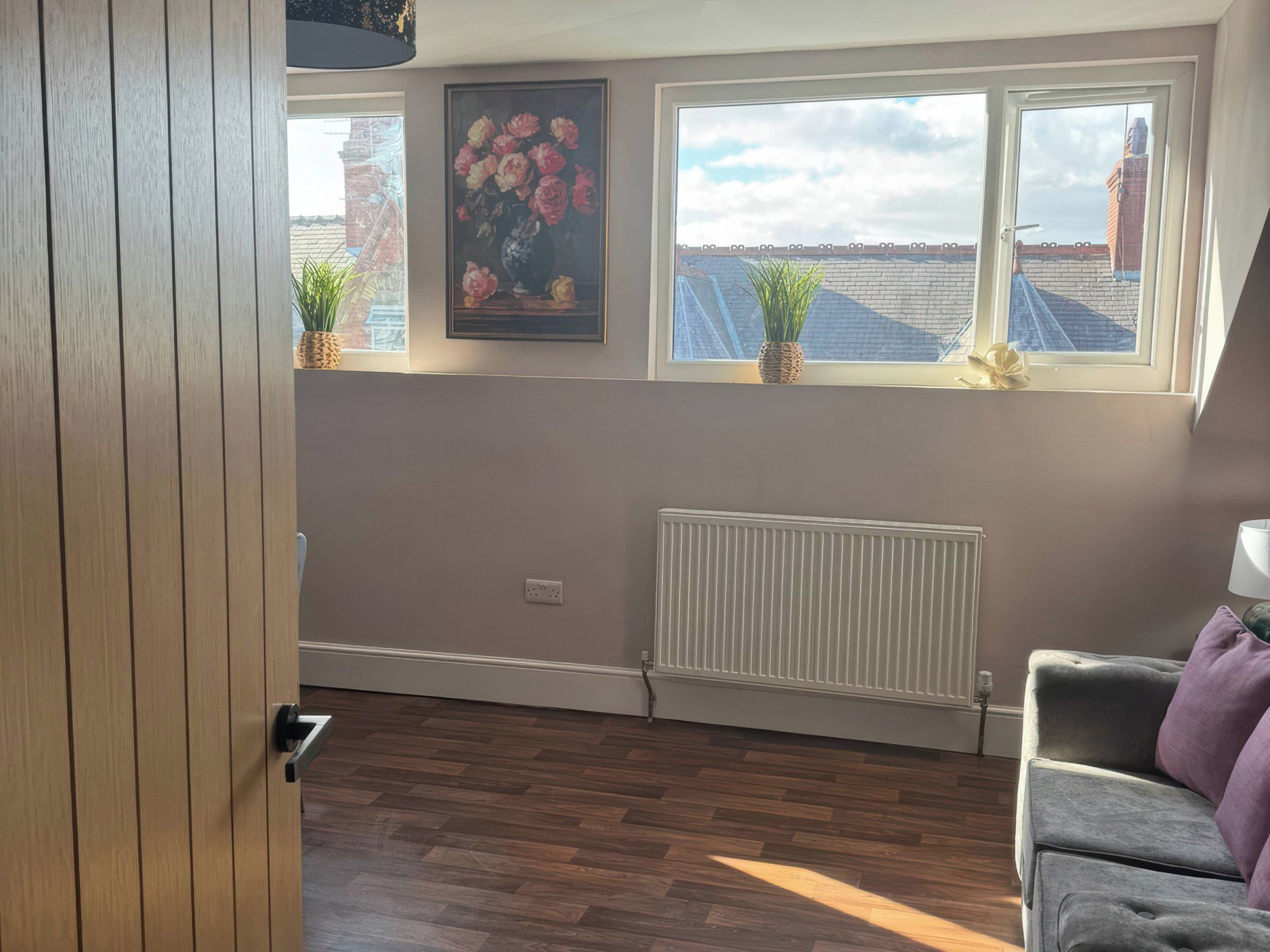 A room with a radiator, two windows with plants, a door, and a view of buildings outside.