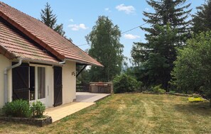 Ein Haus mit Ziegeldach, einer Haustür und einer kleinen Veranda. Ein gepflasterter Weg führt zum Haus, und ein Rasen mit Bäumen erstreckt sich davor.