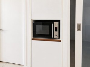 A compact microwave oven installed in a white cabinet with a wooden shelf, located in a room with white walls and a tiled floor. - Brisbane City Views | 2BR Near South Bank & QPAC (South Brisbane)