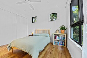 A bedroom with a bed, a nightstand, a bookshelf, and a window with a view of trees.