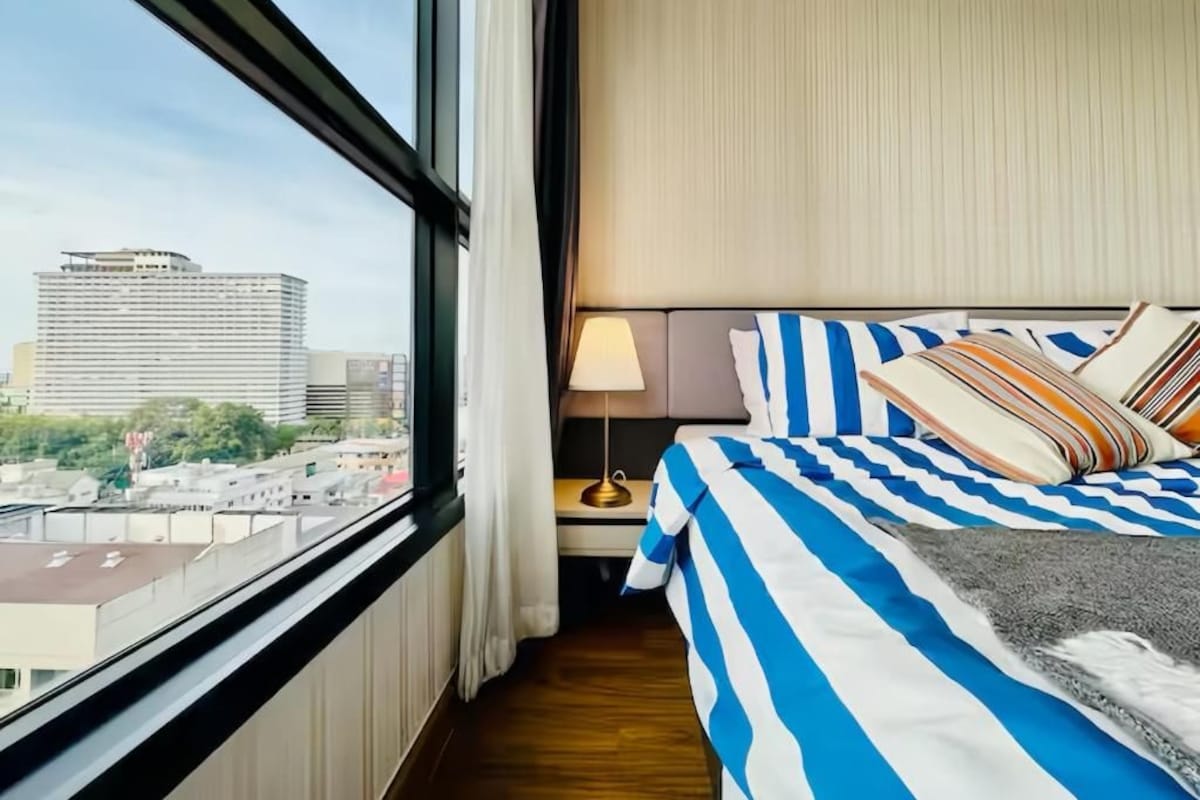 A hotel room with a large window, a bed with striped bedding, a nightstand with a lamp, and a view of a cityscape.