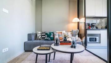 A modern living room with a grey sofa, two round tables, and a chessboard. There are books, a lamp, and a kitchen visible through a glass door.