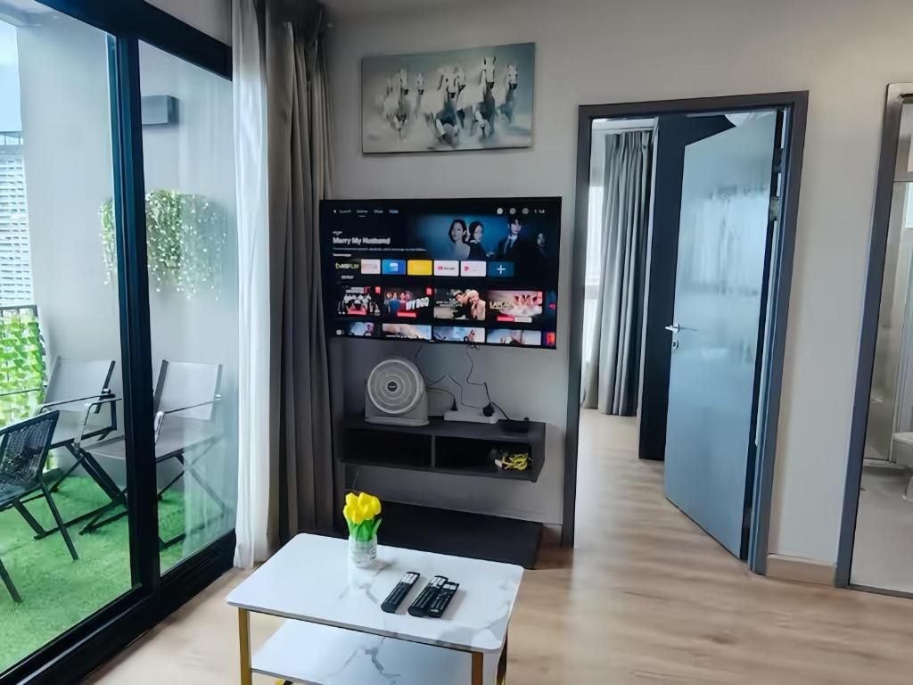 Una sala de estar moderna con una pared de vidrio, un televisor de pantalla plana, una mesa blanca y un jarrón con flores.