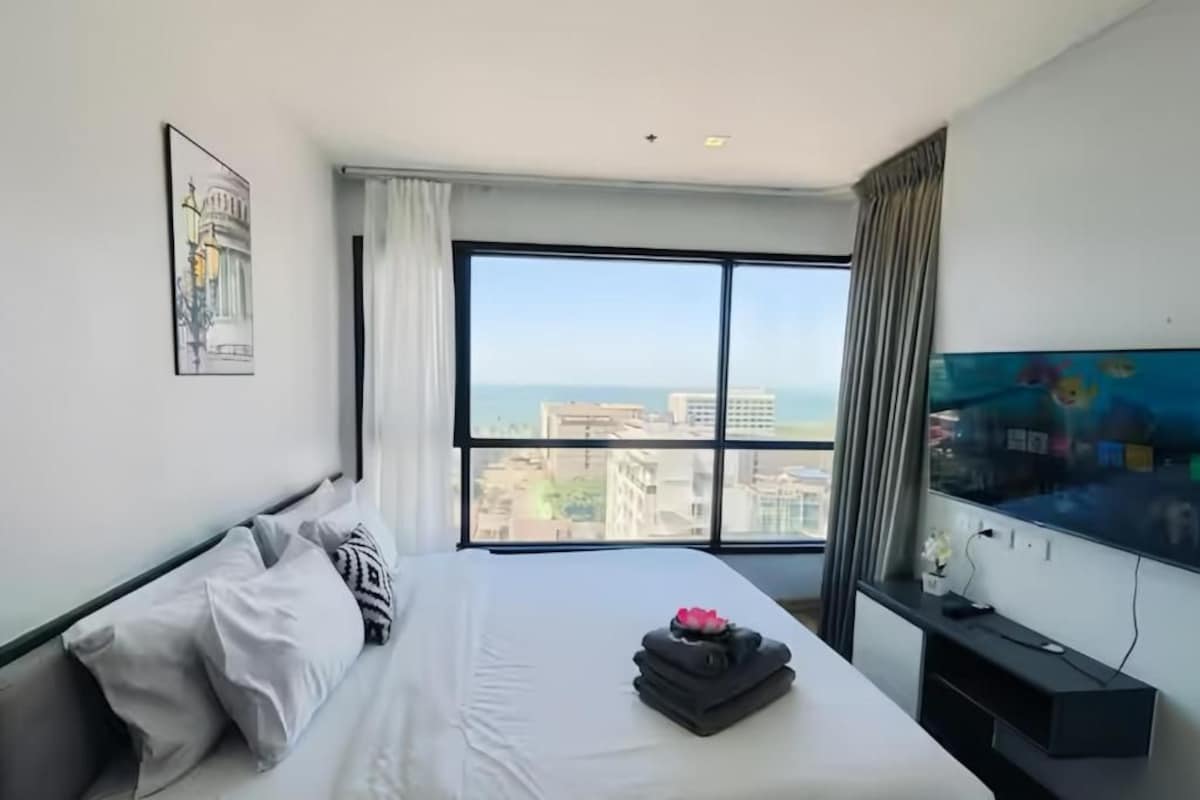 A hotel room with a large window, a bed with white linens, a flat-screen TV, and a painting on the wall.