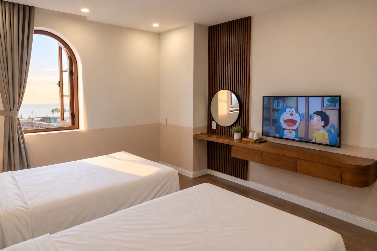 A hotel room with two beds, a TV, a mirror, and a view of the sea.