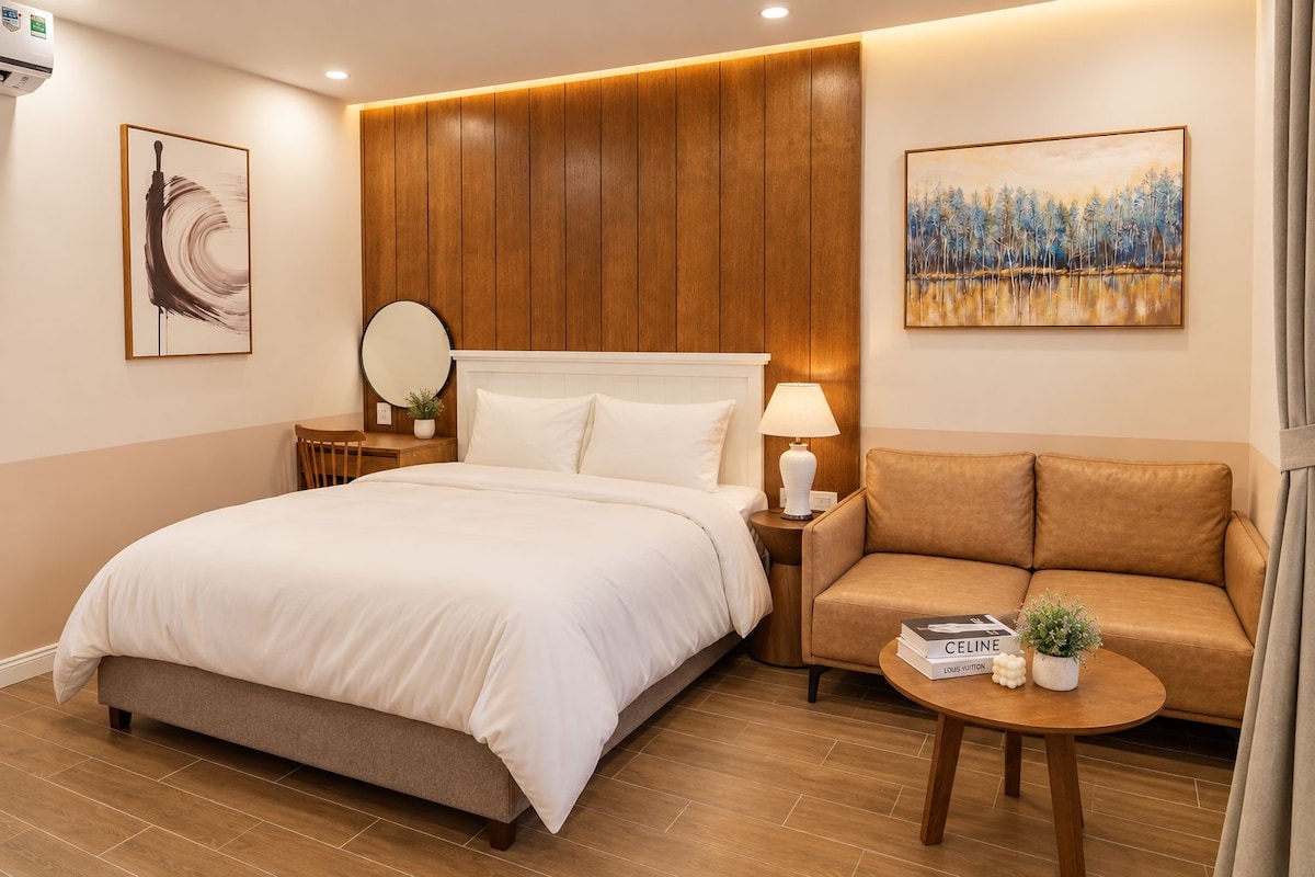 A hotel room with a large bed, a sofa, a round table, a nightstand, a lamp, a mirror, and two paintings on the walls.