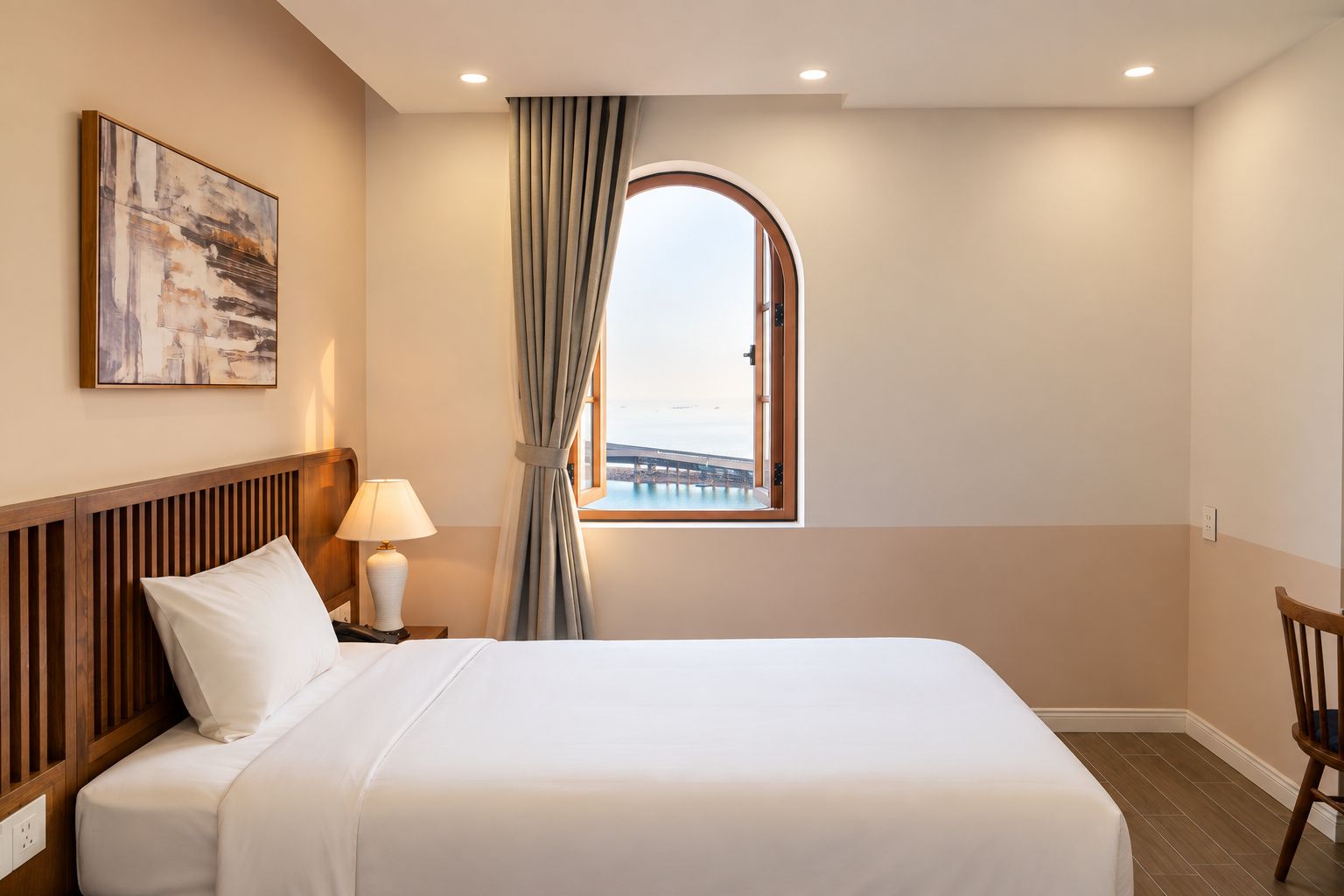 Habitación de hotel con cama, mesita de noche con lámpara, una silla, ventana con vista a un puente y un cuadro en la pared.