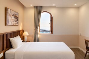 A hotel room with a bed, a nightstand with a lamp, a chair, a window with a view of a bridge, and a painting on the wall. - Palo Santo Phu Quoc - Fireworks & Ocean (Phu Quoc)
