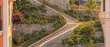 A spiral staircase with stone walls and pink flowers, surrounded by buildings with balconies and shutters.