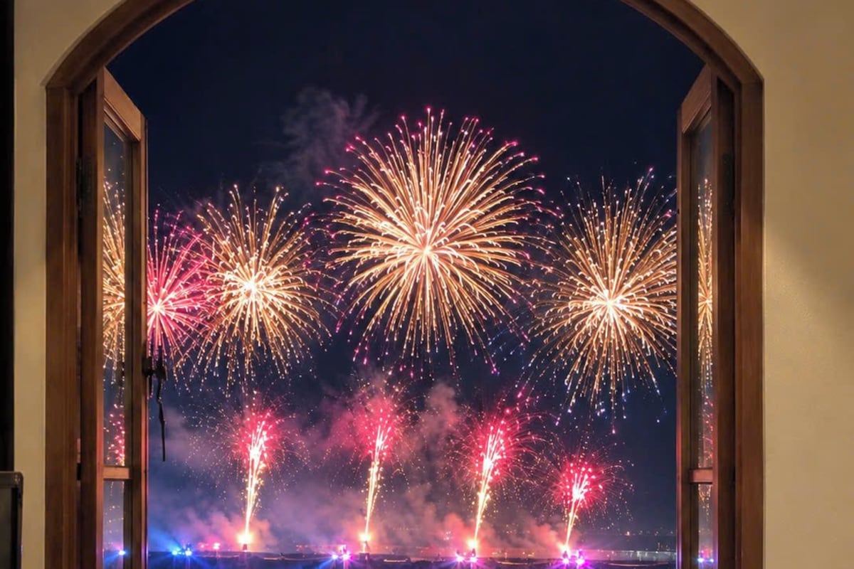 A room with a large arched window overlooking a body of water, with reflections of fireworks.