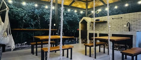 A rustic interior with wooden tables and benches, a hanging swing, and a thatched roof.