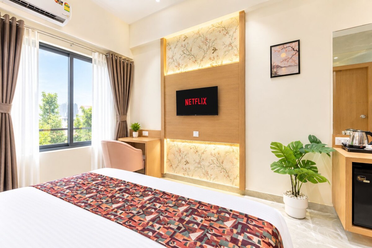 Habitación de hotel con cama, escritorio, televisión montada en la pared, planta en maceta y vistas al exterior.