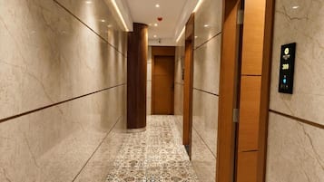 A corridor with marble walls and patterned flooring, leading to an elevator with a digital display.