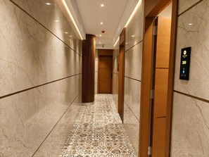 Corredor com paredes de mármore e piso estampado, que leva a um elevador com visor digital.
