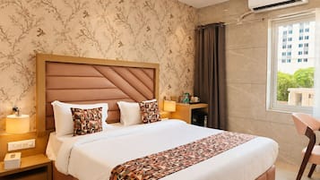 A hotel room with a large bed, a nightstand, a bedside table, an air conditioning unit, a window with curtains, and a view of a building outside.