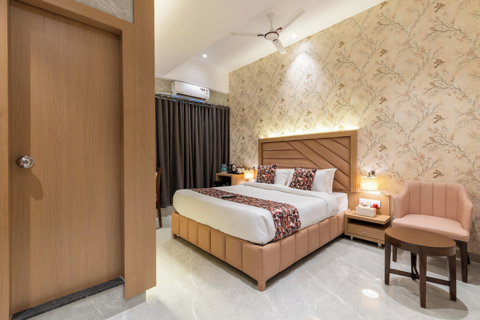 Une chambre d’hôtel comprenant un lit, des tables de chevet, une chaise, un ventilateur de plafond et un mur recouvert d’un papier peint à motifs floraux.