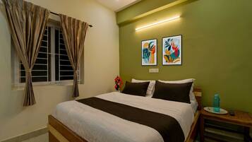 Habitación de hotel con cama, cortinas, mesita de noche, un jarrón de flores y dos obras de arte en la pared.