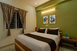 Habitación de hotel con cama, cortinas, mesita de noche, un jarrón de flores y dos obras de arte en la pared.