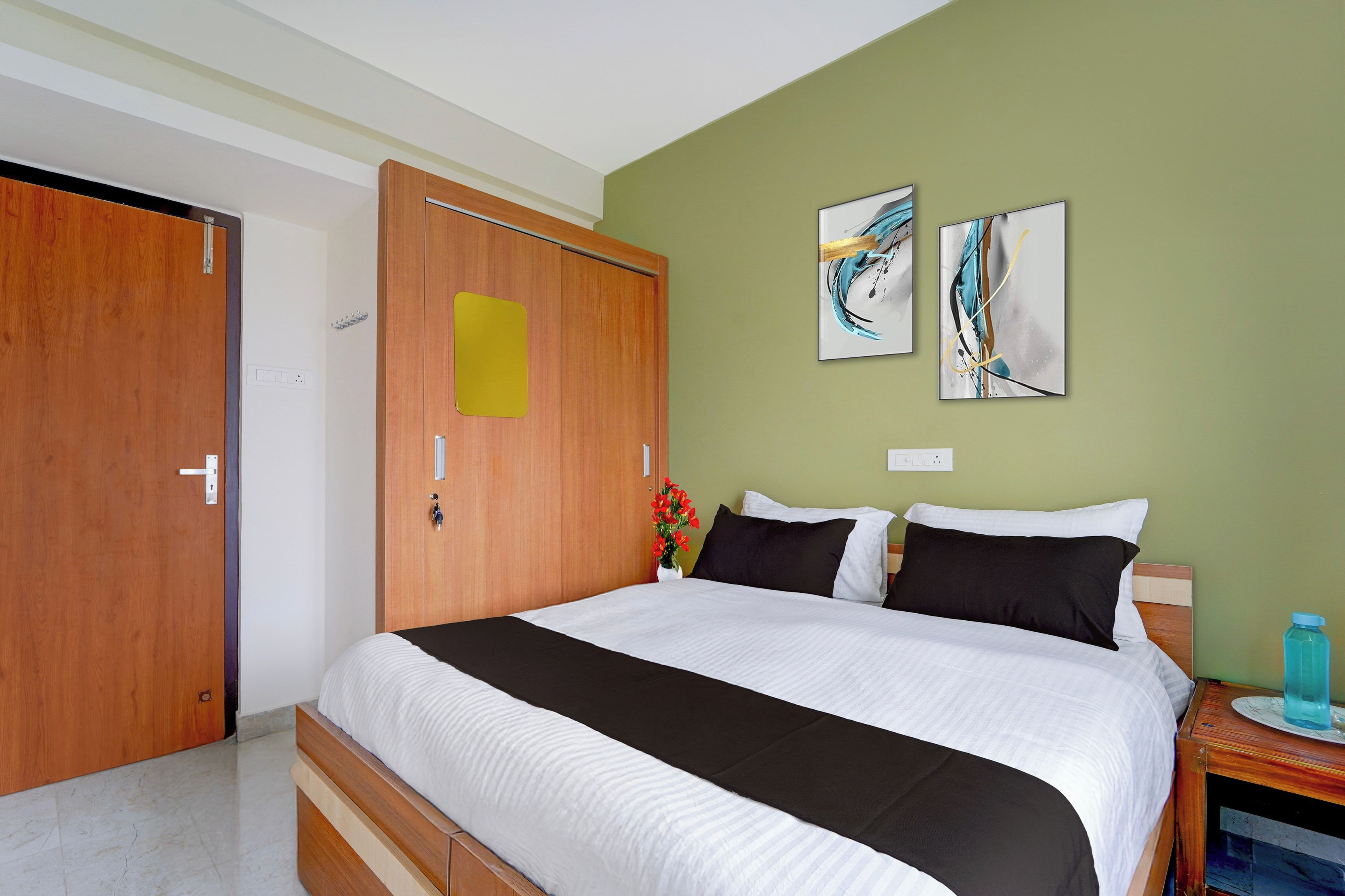 A hotel room with a bed, a wooden wardrobe, a wall with two abstract paintings, and a small bedside table with a water bottle.