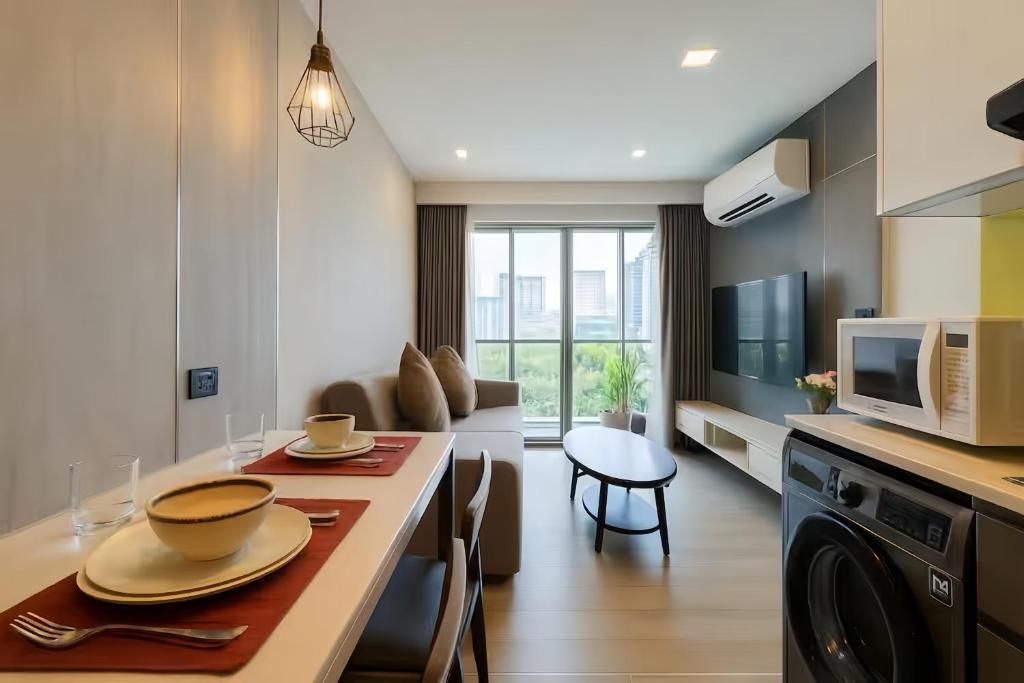 Une cuisine moderne équipée d’un lave-linge, un coin repas avec une table dressée pour deux personnes et un salon avec un canapé et un téléviseur.