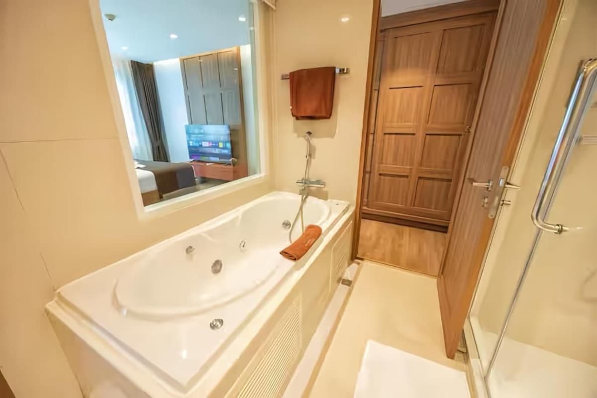 Un baño con una bañera grande, una puerta de madera y un espejo que refleja una habitación con cama y televisor.